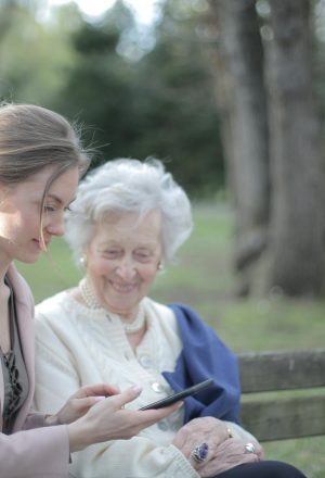 Side view of smiling adult female helping aged mom in using of mobile phone while sitting together in park